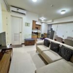 A bright, modern apartment interior with a sectional sofa, dining area, and kitchen.