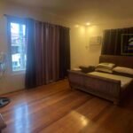Bedroom with hardwood floors, wicker bed frame, curtains, and a standing fan.