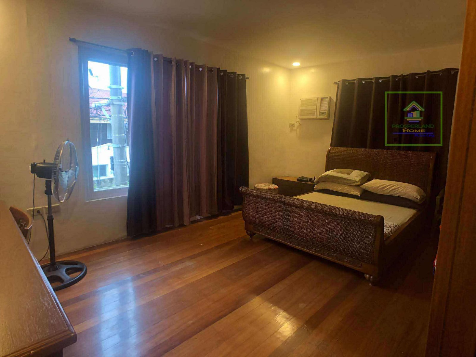 Bedroom with hardwood floors, wicker bed frame, curtains, and a standing fan.