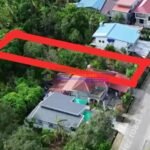 Aerial view of houses and a vacant lot outlined in red, surrounded by lush greenery and a road.