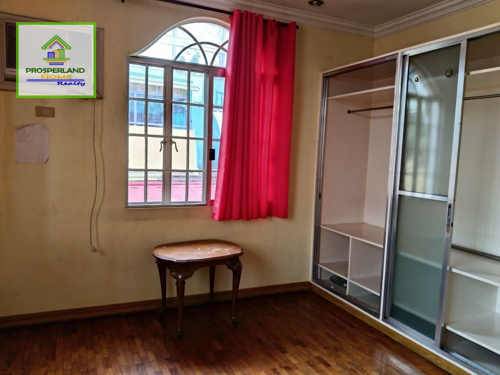 Bedroom with wood floors, a window with red curtains, a small table, and a sliding-door closet.