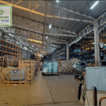 An interior view of a large warehouse filled with materials, including glass and wooden crates, under a high, exposed-beam ceiling.
