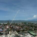 Construction site with crane, buildings, and a lake in the background under a cloudy sky.