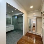 Interior shot of a bathroom and closet area with light walls, tile, wood floors, vanity, glass shower, and mirrored closet doors.