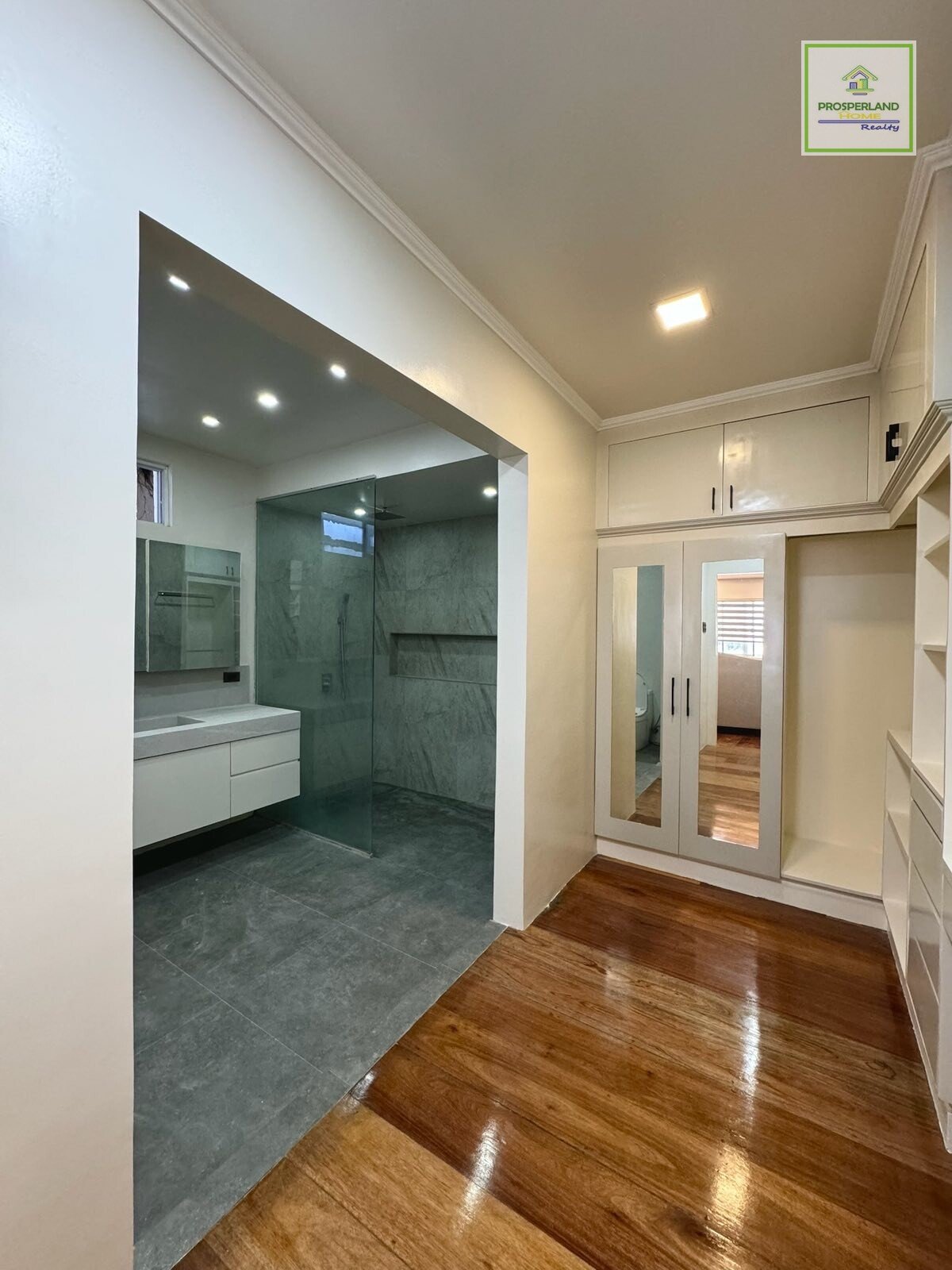 Interior shot of a bathroom and closet area with light walls, tile, wood floors, vanity, glass shower, and mirrored closet doors.