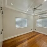 Bright, empty room with hardwood floors, two windows with blinds, a ceiling fan, and Prosperland Realty logo.