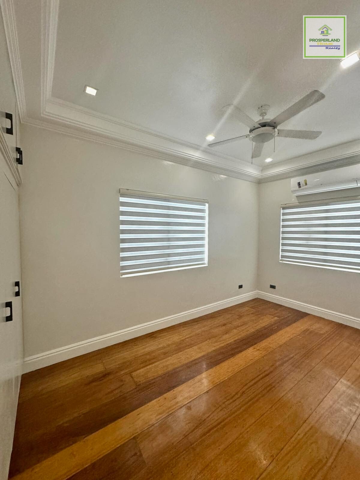 Bright, empty room with hardwood floors, two windows with blinds, a ceiling fan, and Prosperland Realty logo.