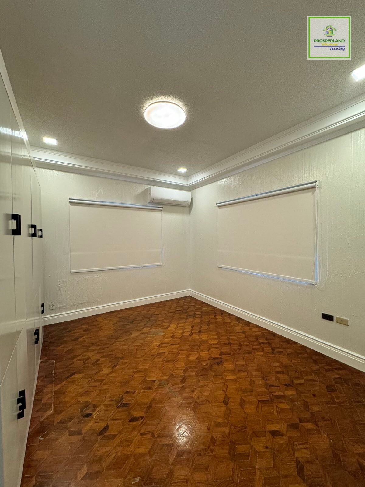 An empty room with parquet floors and two windows with white blinds.