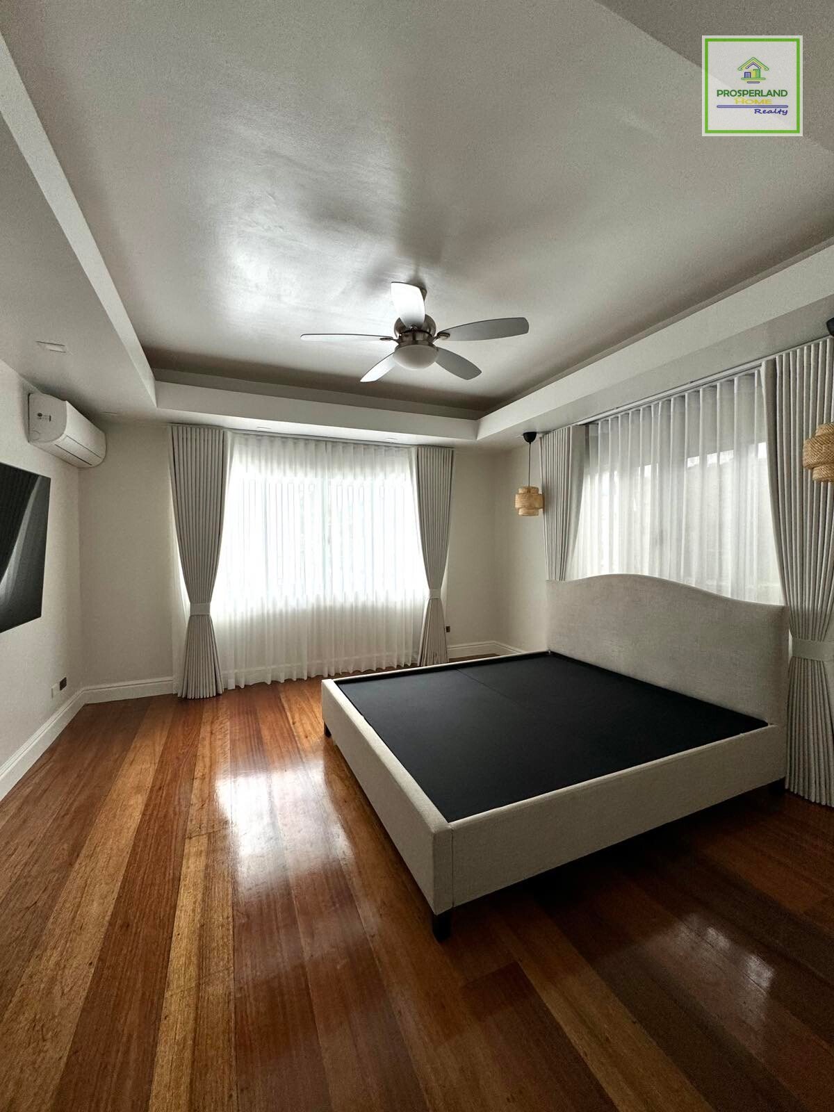 Bright bedroom with wood floors, a black mattress on a beige platform bed, sheer curtains, and a ceiling fan.