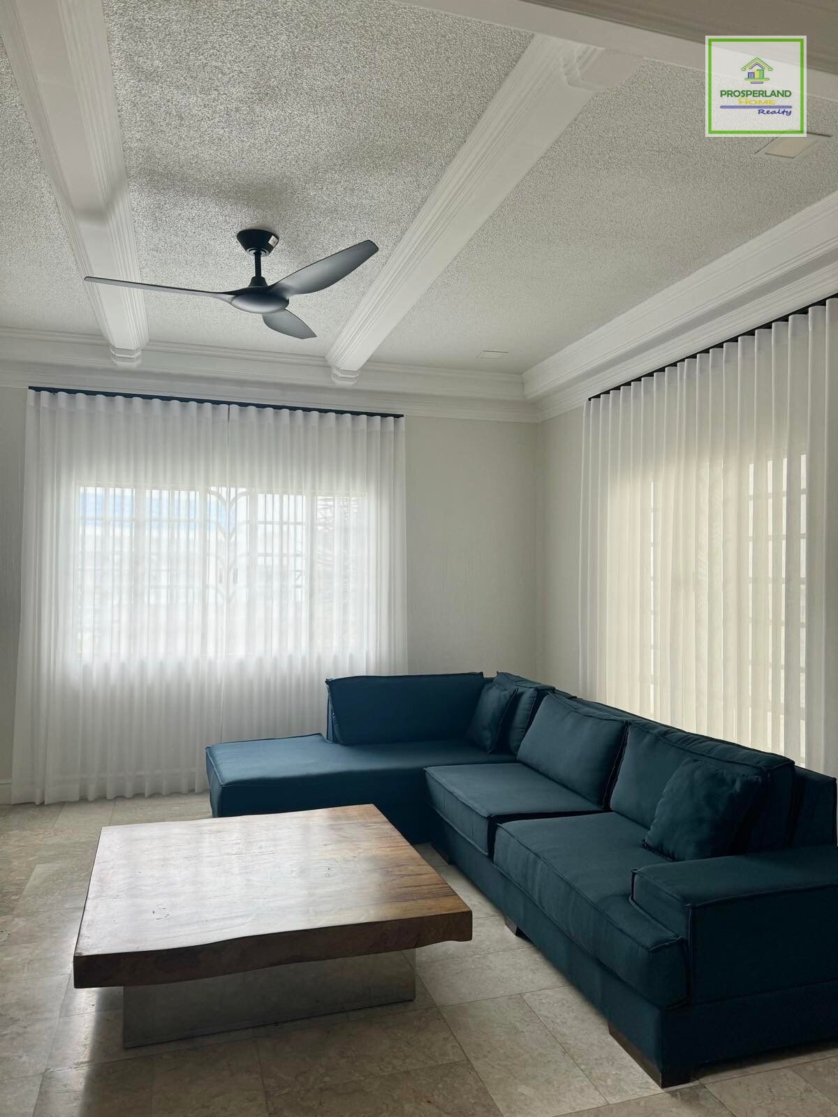 A modern living room with a blue sectional sofa, wood coffee table, and white sheer curtains.