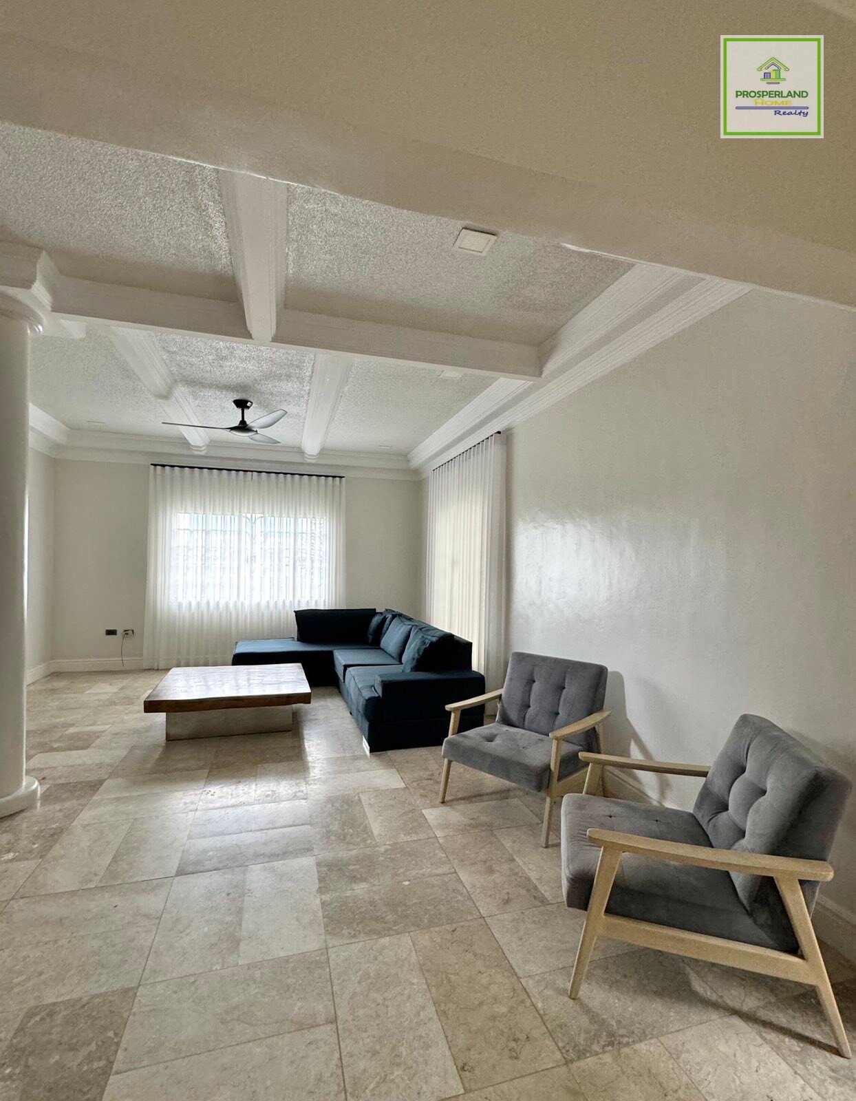 A bright living room with a dark blue sectional, two gray armchairs, and beige tile flooring.