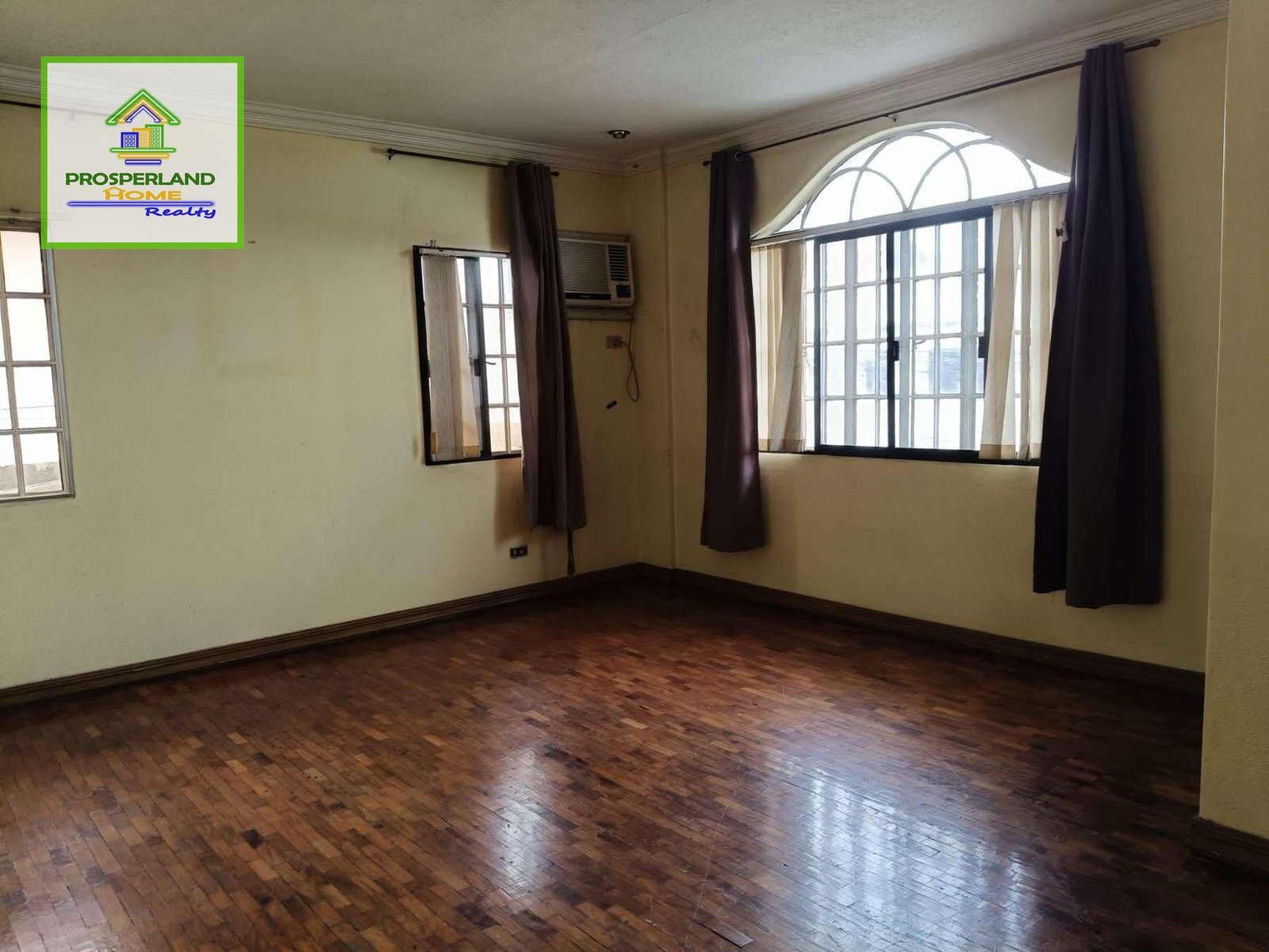 An empty room with hardwood floors, windows with curtains, and tan walls.