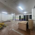 A mostly empty room with fluorescent lighting, wooden flooring, and some scattered debris.