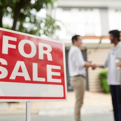 A "For Sale" sign is in focus, with a blurred couple and real estate agent shaking hands in front of a house.