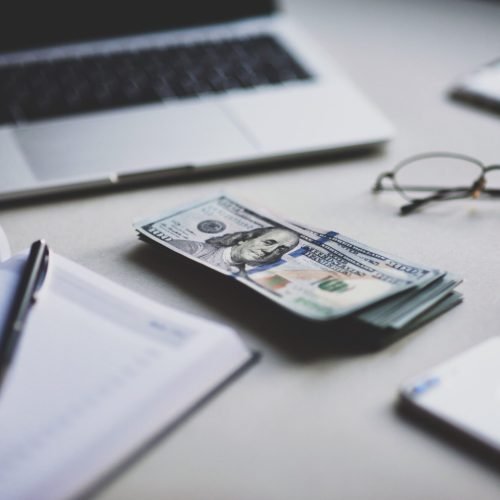 Flat lay of laptop, notebook, pen, money, glasses, and phone on a desk.