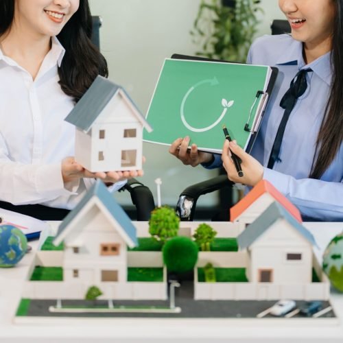 Two women discuss sustainable housing with model houses, wind turbines, and a recycle symbol.